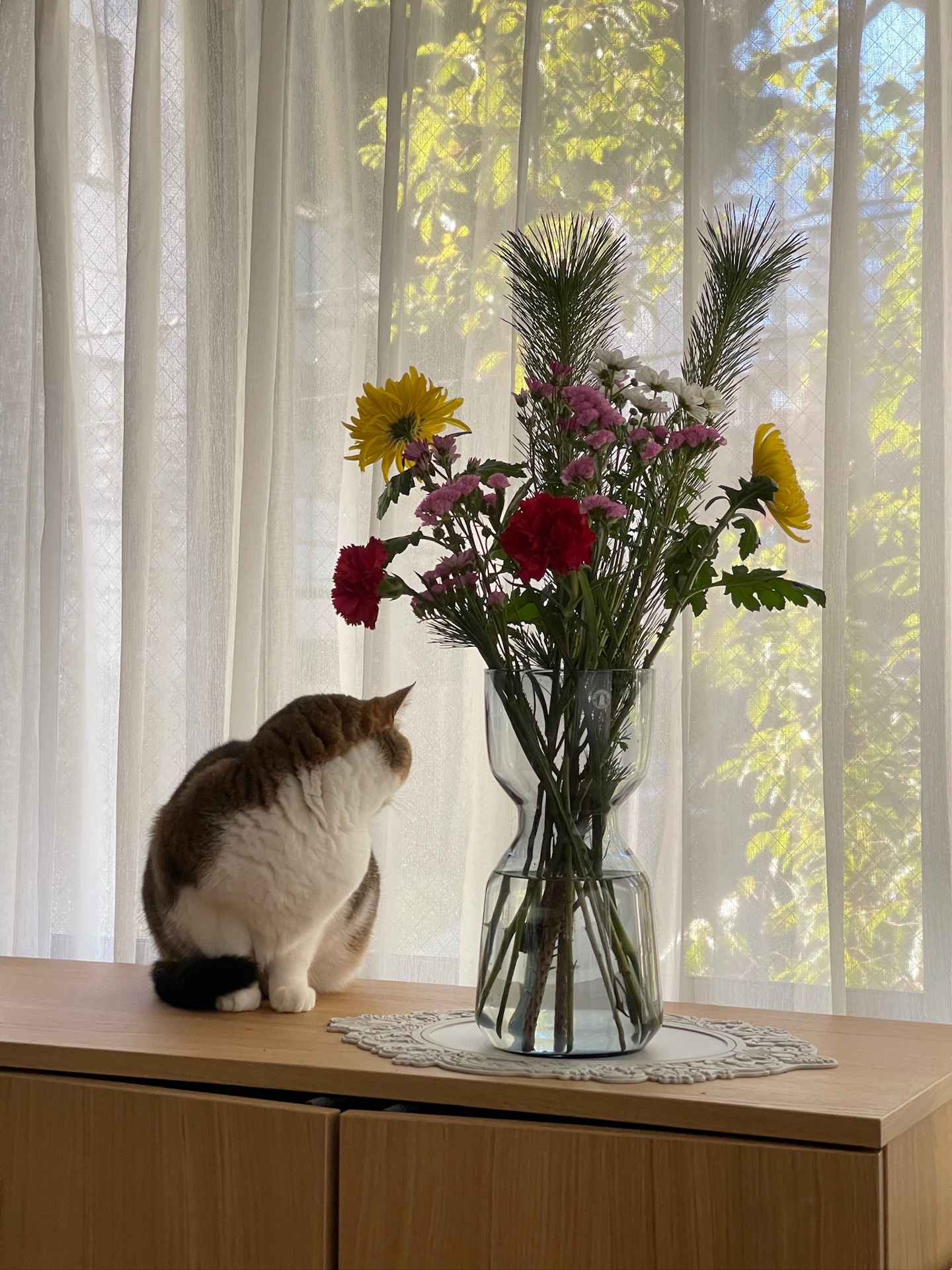カーテン越しの緑を背景にした花器と、窓辺のキャビネット